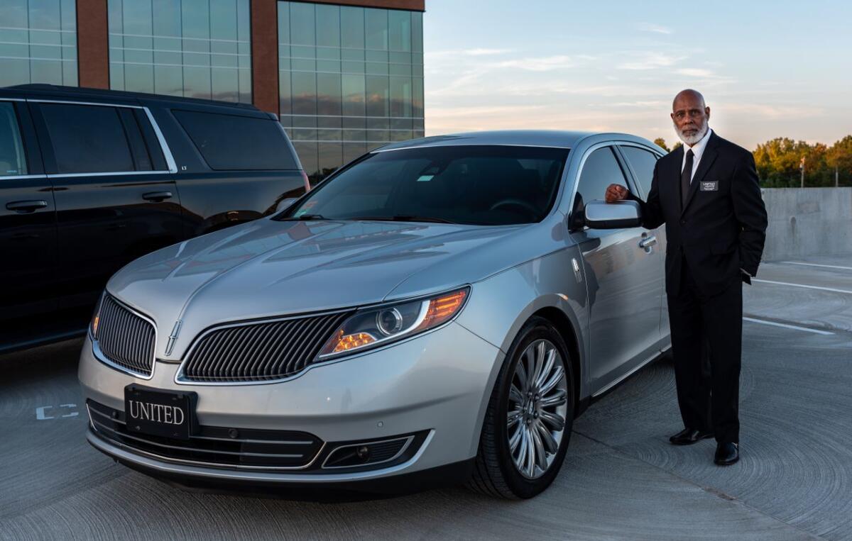 services-01 Professional chauffeur in a suit standing next to a luxury car in outdoor setting.